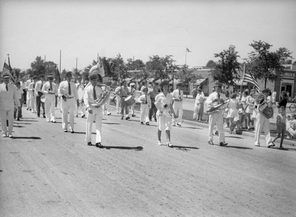 A marching band in full regalia in a Fourth of July parade. They are walking in the street, spectators are along the grass near the curb, and storefronts are visible in the background. Men, women and children are carrying instruments, including tuba, french horn, drum, trombone and saxophone.