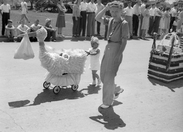 A young girl pushes a baby carriage along a street in a Fourth of July parade. The carriage is decorated like a stork with a bundle tied to its beak. A woman walks beside the young girl, and spectators line the side of the street along the curb.