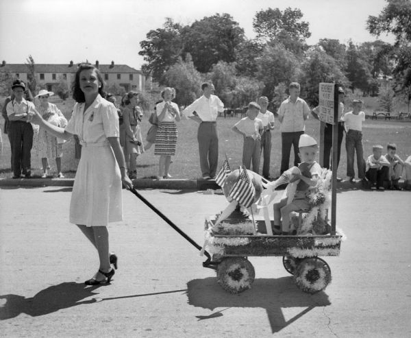 Side view of a woman pulling a little boy in a decorated coaster wagon in a Fourth of July parade. The boy is holding a bottle of milk with white streamers coming from it which are flowing onto a globe of the world sitting in front of him. A sign behind the boy reads "REMEMBER MILK OF HUMAN KINDNESS FEEDS THE WORLD ALSO." Spectators line the curb of what appears to be a park in the background.