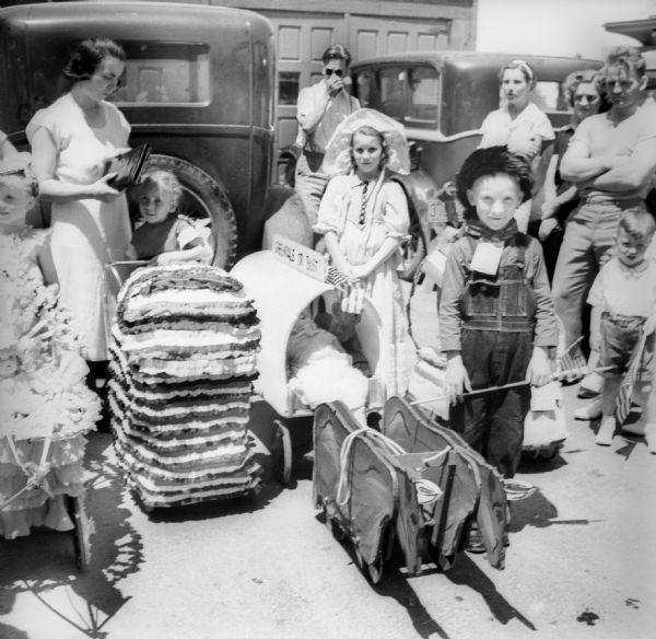 Children in costumes stand with adults near decorated baby carriages and coaster wagons before a Fourth of July parade. One of the wagons is covered and has a sign that says "Greendale or Bust" with a pair of wooden horses to pull it. In the background are two automobiles.