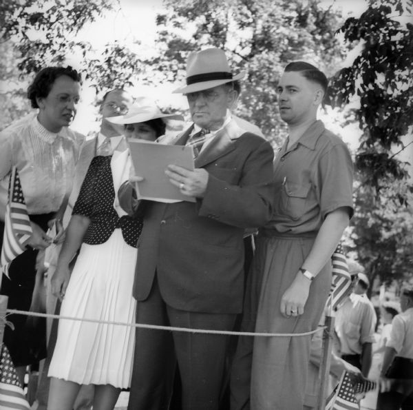 Men and women standing on a roped off platform before a Fourth of July parade. The man wearing the hat and eyeglasses is potentially the grand marshal of the parade.