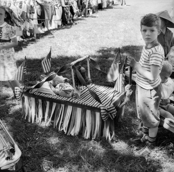 A small boy is standing next to a baby in a decorated coaster wagon before a Fourth of July parade. A large crowd is standing on the grass behind them, and many people are holding flags.