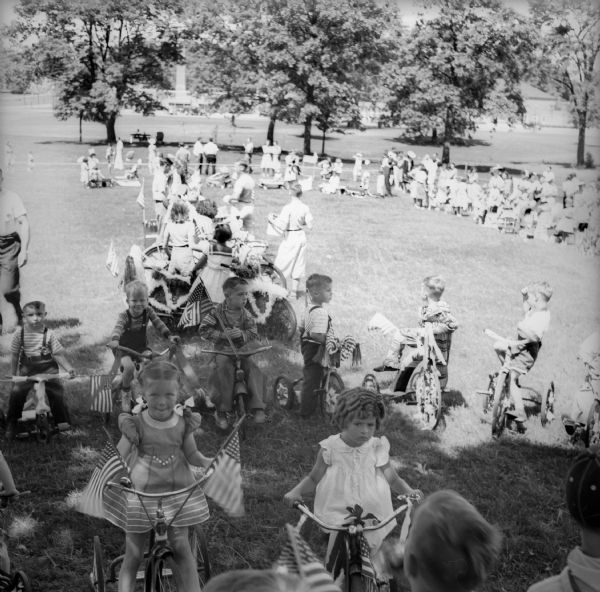 A crowd of children are lines up in a park with bicycles and tricycles decorated with streamers and tissue paper before a Fourth of July parade.