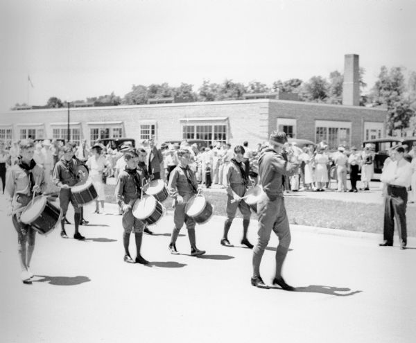 A group of Boy Scouts, led by the Scout Leader, playing drums in a Fourth of July parade. They are walking down a street and a crowd of spectators is in the background near a building.