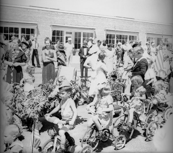Children riding decorated tricycles and baby carriages in a fourth of July parade with adults lining the street behind them.