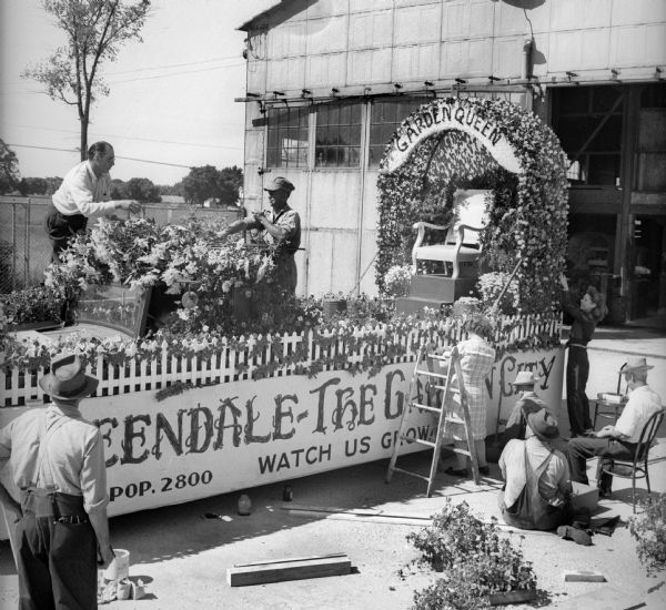 Group of people decorating the Garden Queen float for a Fourth of July parade near a large garage. The float reads "Greendale-The Garden City," "Pop. 2800" and "Watch Us Grow." The float is a car or truck hidden by a picket fence decorated with leaves and flowers, and there is a chair under an arbor at the rear of the float with a "Garden Queen" sign.