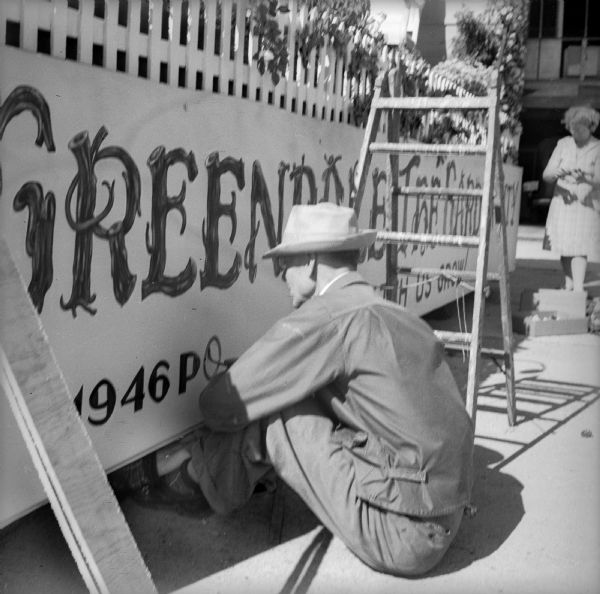Jack Rickert is painting words "1946 Pop. 2800" on the side of a Fourth of July Garden Queen Float. Above the painted sign the float is decorated with a picket fence and flowers. There is a woman in the background and a large garage behind her.