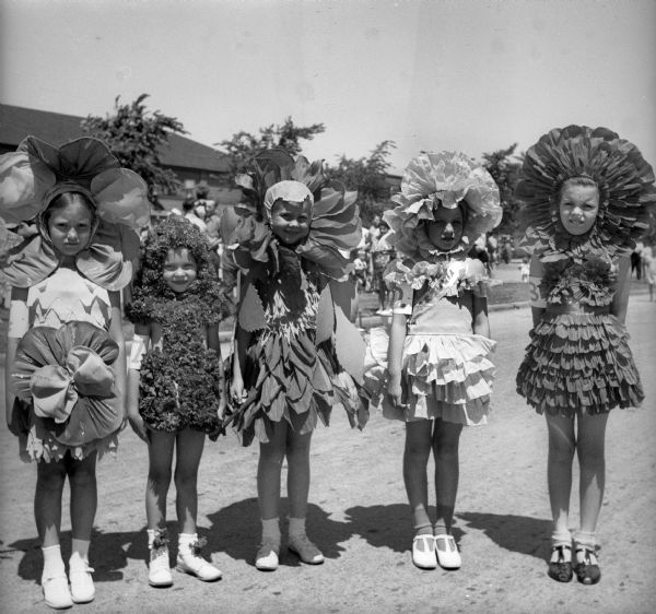 Five girls dressed as flowers, probably as part of a Fourth of July "Garden Queen" pageant. The girls are standing in a row and each of the girls has a number around her arm. One girl has a sash that reads: "Petunia."