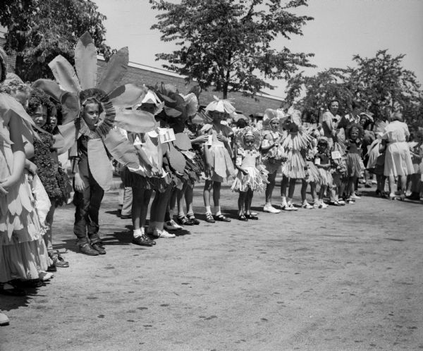 A line of girls dressed as flowers, probably as part of a Fourth of July "Garden Queen" pageant. The girls are standing in a long line and each of the girls has a number on her arm or costume.
