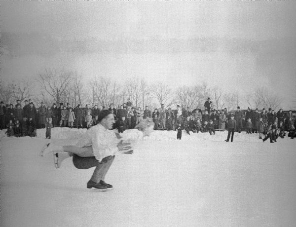 A couple, most likely Gerda Wank with Joe Puccetti, in the middle of an ice skating exhibition. The man is holding the woman almost parallel to the ice while crouching. A crowd of children and adults is watching from a snowbank in the background.