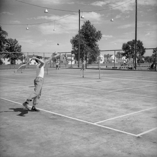 A young boy is in mid-swing playing tennis on a tennis court. Other tennis players, houses and a park are visible in the background.