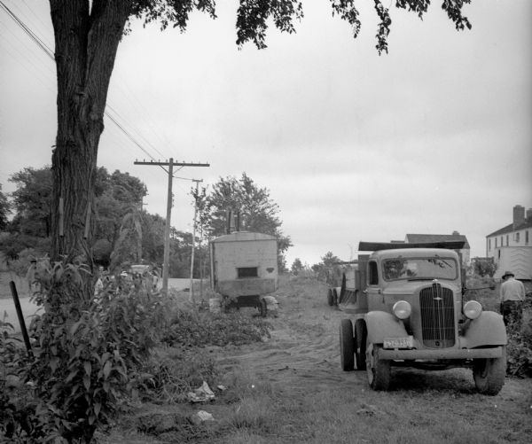 A crane and a truck in a clearing between a road lined with telephone poles and a row of houses. On the right near the truck a man is working. The license plate of the truck reads "US 52-993." There are a man and boy on the road on the left, as well as an automobile.