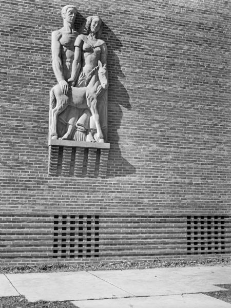 Stone carving of a farm couple with a foal installed on the side of a brick building. Carved by Alonzo Hauser in 1938.