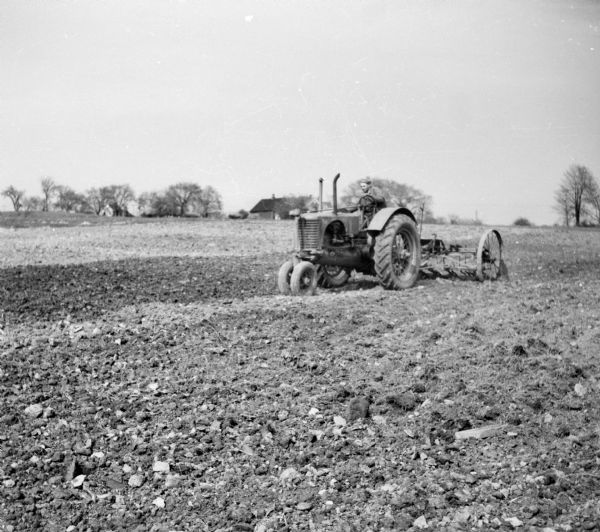 A man tilling soil on a farm with a tractor. Houses and trees are visible in the background.