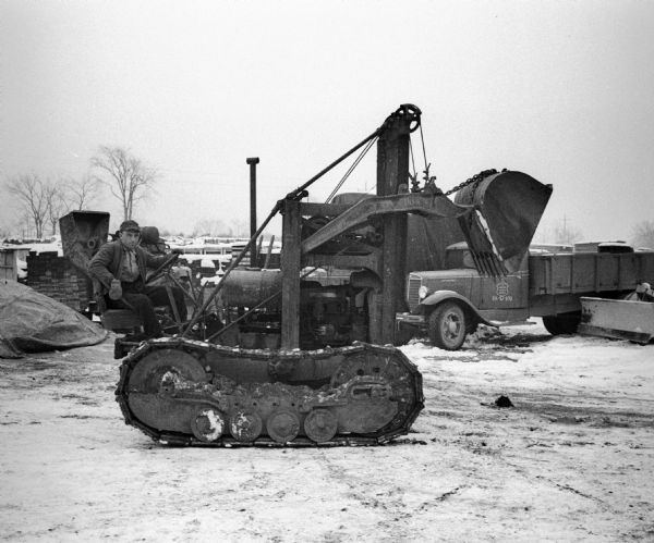 A man is sitting at the wheel of a gas powered bulldozer. In the background are equipment, supplies, and a truck with the "US Dept of Agriculture / Farm Security Administration" logo on its driver-side door. Snow is on the ground.