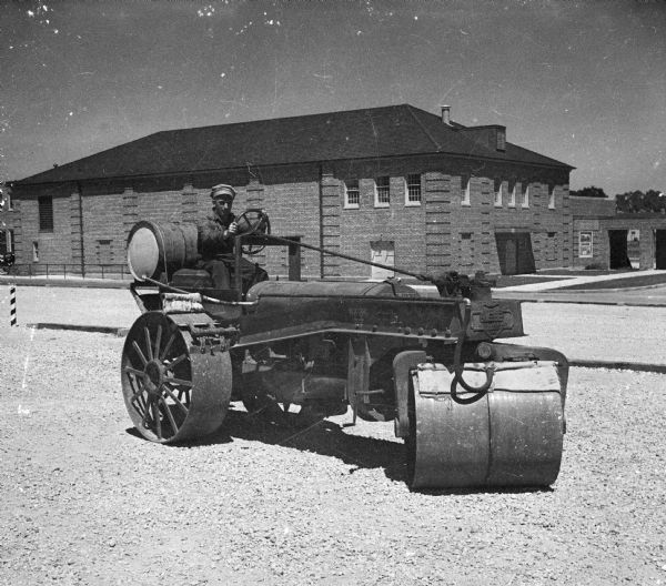 A man is sitting at the wheel of a long steamroller parked in a gravel lot with a brick building in the background.