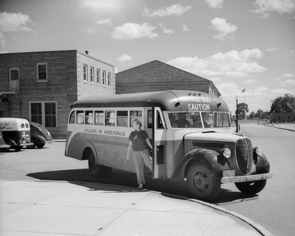 A teacher waiting for students in the doorway of a school bus parked in the driveway near a brick building. A driver and other adults are visible in the interior of the school bus which has "Village of Greendale" written on the side. In the far right background is a flagpole in a park.
