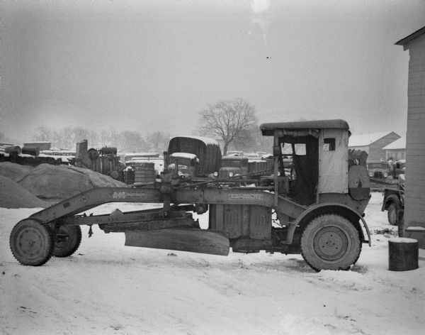 A grader manufactured by The Allis-Chalmers Manufacturing Co. sits in a lot. Trucks and equipment are parked in the background near industrial buildings and snow is on the ground.