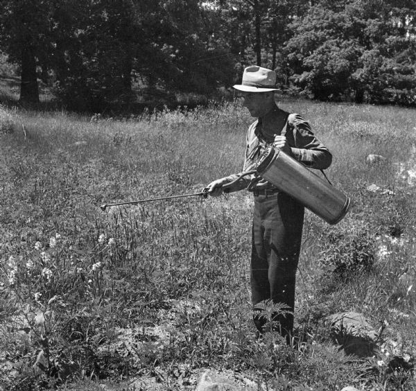 A man wearing a hat sprays liquid from a canister hanging from his shoulder designed to kill mosquitoes. He is in a field and trees are in the background.