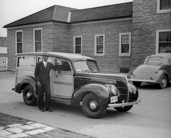 A man, most likely a driver or chauffeur wearing a cap, leans on a Ford automobile in a parking lot. A brick building and another car are visible in the background. The passenger side door of the car has the seal of the "US Dept of Agriculture / Farm Security Administration."
