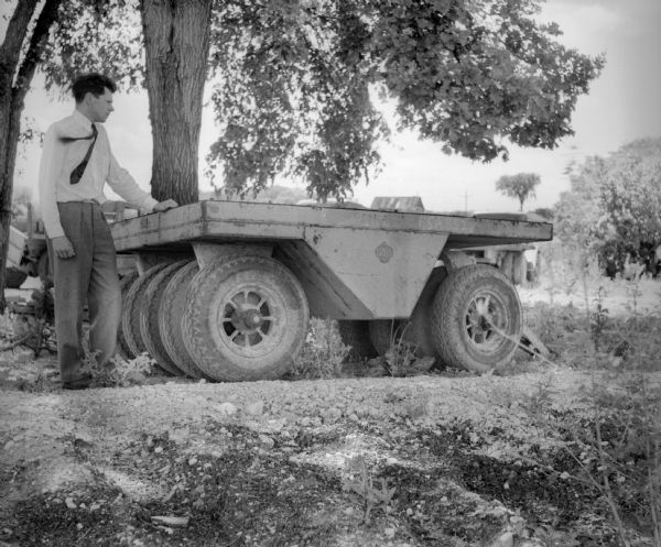 A man wearing a tie stands next to an industrial trailer used for clearing of land. The side of the trailer reads "BROS" and there are other vehicles in the background behind trees.