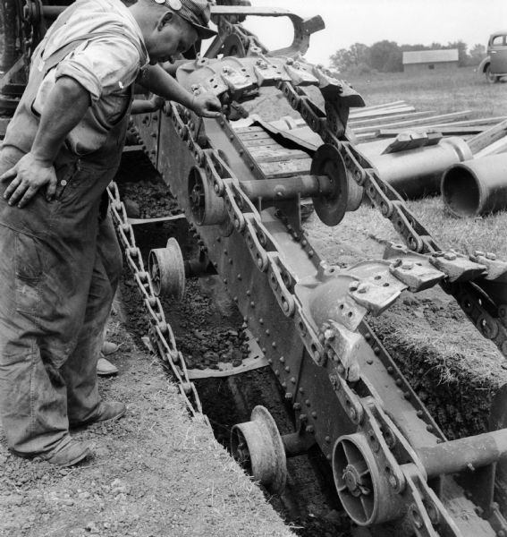 A man in a hat and overalls leans over a trench or ditch digger, looking down into the excavated area. Pipes, an automobile, and a building are in the background.