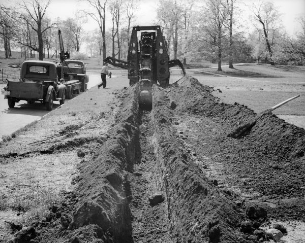 Rear view of a trench dug by a ditch digger. A man walks towards the ditch digger from two trucks, one fitted with a crane-like attachment, which are parked on the side of a road on the left. In the background is an open area with trees, perhaps a park.