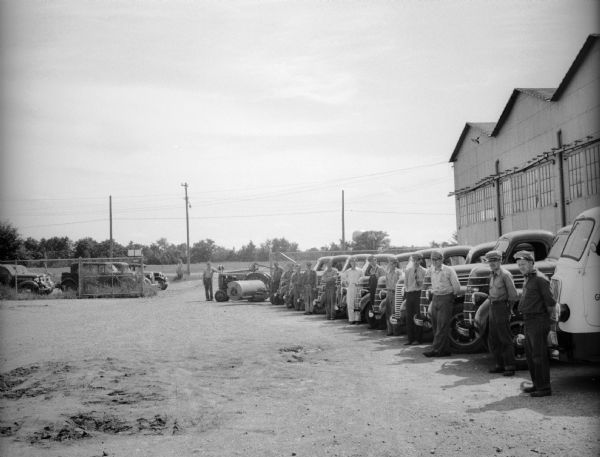 Thirteen men, all wearing hats, standing in a row in front of trucks and a tractor in a lot near an industrial building and chain link fence. Probably service vehicles associated with the US Dept of Agriculture / Farm Security Administration.