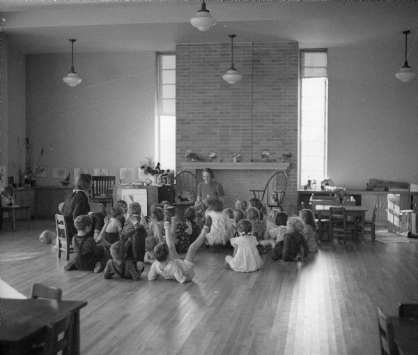 A school teacher reads to elementary school children, who are sitting on the floor in a large, open classroom.