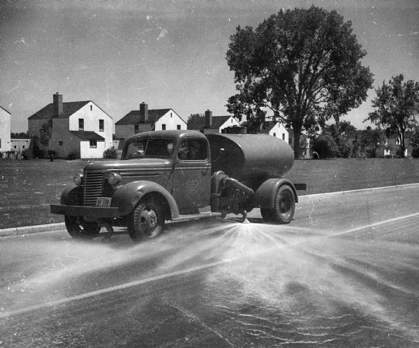 A street cleaning truck spraying water on a suburban street with houses in the background. The text on the driver's side door probably reads "US Dept. of Agriculture / Farm Security Administration."