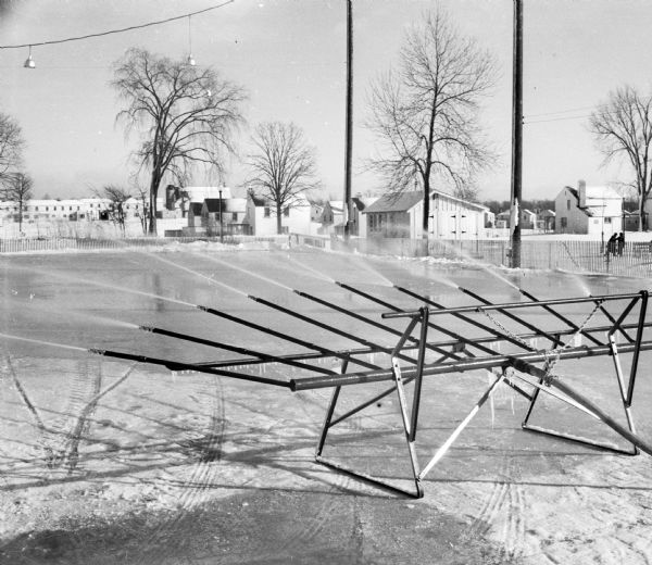 A device spraying water to create an ice rink. The rink was 115 x 200 ft. and was opened sometime during Christmas week 1938. There are houses and pedestrians in the background.