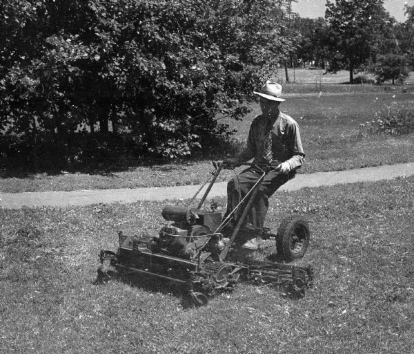 A man wearing a hat, shirt, and tie is driving a gas-powered riding lawnmower.