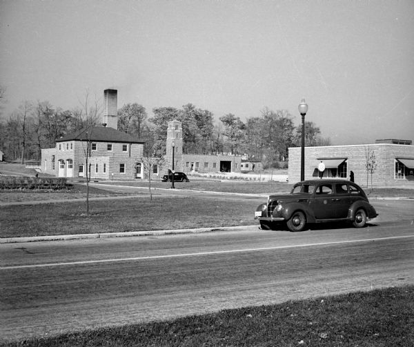 View across road of car parked on the side of the road. In the background are brick buildings, including (from left to right) the Central Heating Plant, the Fire Station, the Police Station and the Public Works Building.