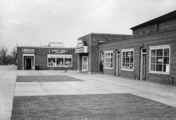 A woman and child in front of brick commercial storefronts. Signs read, from left to right "Drugs Prescriptions," "Soda Grill Luncheonette" and "Greendale Theater, Air Conditioned For Comfort, New Show Every Wed Fri Sat."