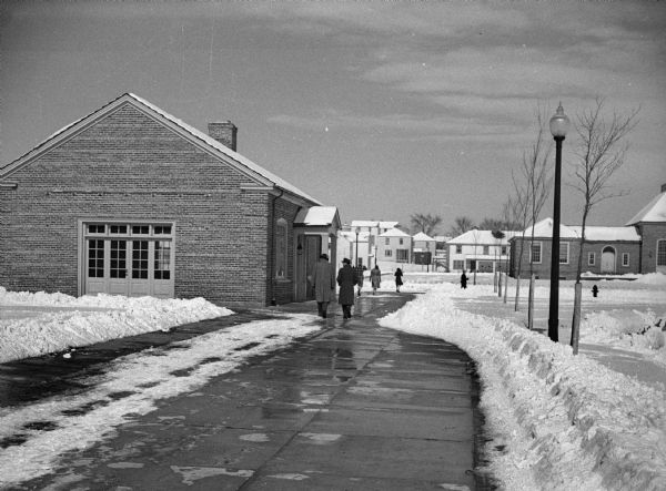 Men and women walking on sidewalk in front of a brick commercial building. The Greendale Village Hall is in the background and snow is on the ground.
