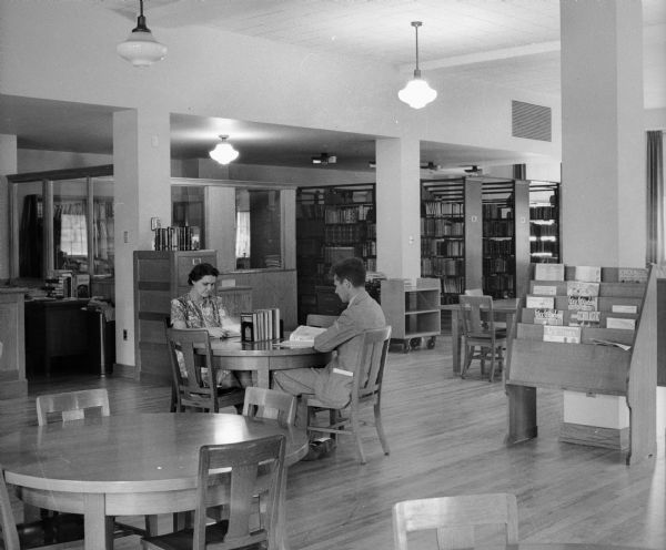 A woman and man sit at a round table in a public library reading. An office cubicle and magazine rack are visible in the background. When it opened in 1938, the Library had a seating capacity of 72 and a 5600 volume capacity, though it only had 2000 volumes at the time.