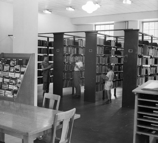 Three boys in a public library looking for books in the main stacks. A table and magazine rack are in the foreground. When it opened in 1938, the Library had a seating capacity of 72 and a 5600 volume capacity, though it only had 2000 volumes at the time.