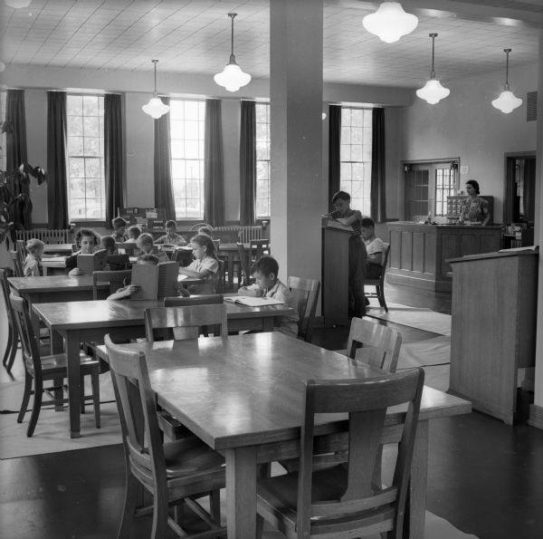 A group of children sit at tables in a library reading books and magazines while a librarian looks on from her circulation desk. When it opened in 1938, the Library had a seating capacity of 72 and a 5600 volume capacity, though it only had 2000 volumes at the time.
