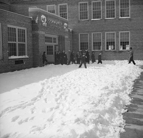 A group of children exiting the brick Community Building. Two sculptures flank the sign above the entrance. There is a thick layer of snow on the ground and the children are wearing coats, hats and mittens. There is a man standing on the sidewalk on the right.