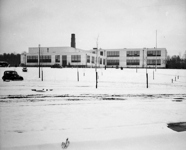 View across snow-covered lawn or field of a school building with a smokestack. Automobiles are parked nearby.