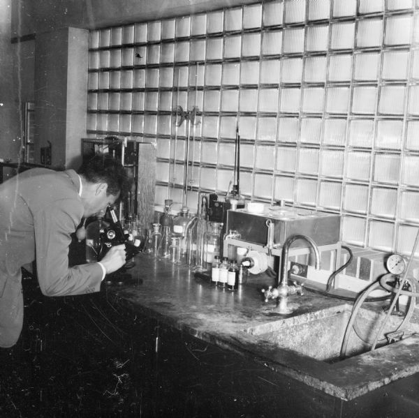 A scientist leans over a microscope in the sewage treatment plant laboratory. Glass beakers and other tools and equipment are sitting on the counter.