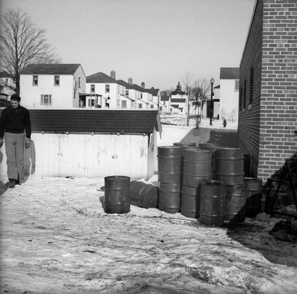 A pile of metal drums sits in the snow next to corner of brick building. One of the drums has the words "Hello Neighbor" painted on it. Residential housing is visible in the background, and a young man stands on the left.