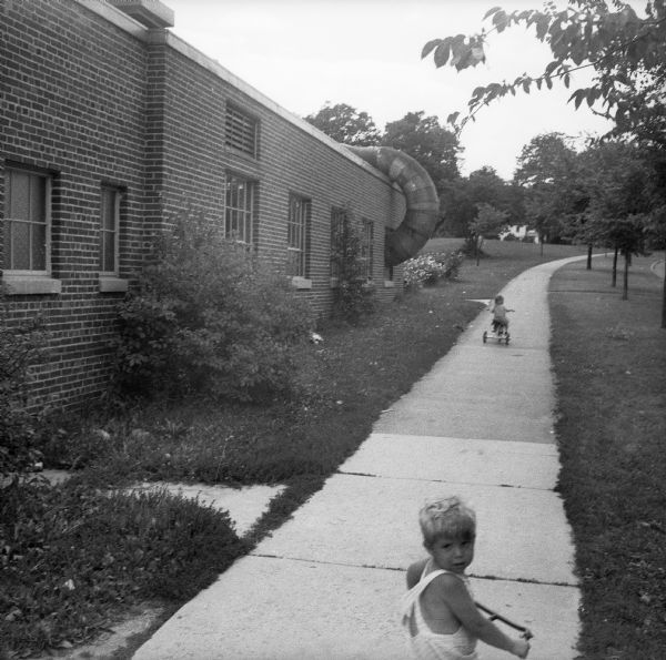 Two children ride tricycles on sidewalk along the side of a brick building. There is a large pipe winding from the roof into one of the windows.