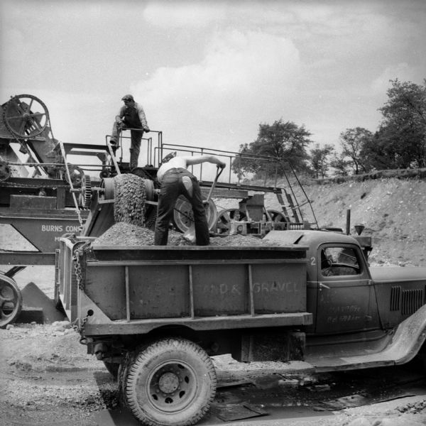 A man is standing on a walkway on top of a mechanical conveyor belt machine in a quarry, loading a truck with sand and gravel. A man with a shovel is standing in the truck to redistribute the gravel evenly. The conveyor machine has the text "Burns Cons[obscured]" written on it, and the truck has the text "Washed Sand & Gravel" written on its side.