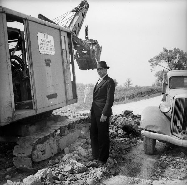 A man wearing a suit and a hat stands on a mound of dirt in a road with an automobile on his left and a steam shovel on his right. The steam shovel has a sign on its side reading: "General Excavator Cos. / Invincible / Marion, Ohio U.S.A."