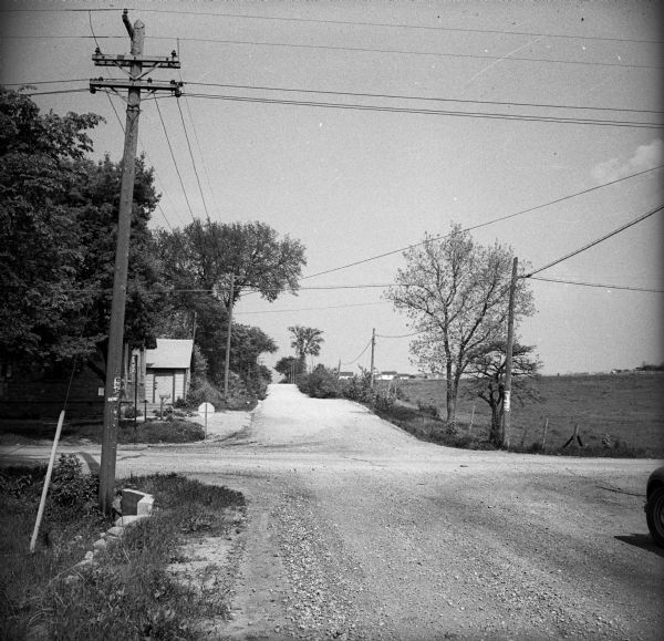 Intersection of two gravel roads. Telephone poles, some with posters stapled to them, line the sides of the road, and residential housing and fields are visible in the distance.