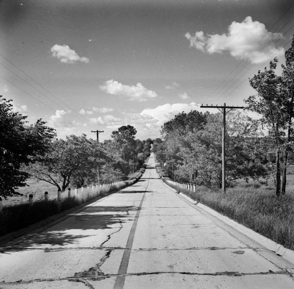 View looking down two-lane road to the horizon. Telephone poles, power lines, and trees are on either side, with fields behind.