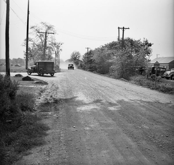 Two cars driving on a gravel road lined with telephone poles or power lines. On the right beyond a stop sign, cars are parked behind a fence. More cars are parked in a field on the left.