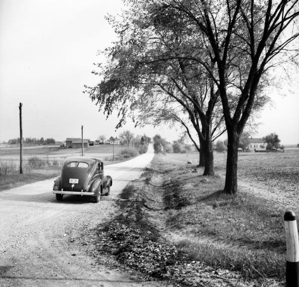 A car is parked on a gravel road that stretches to the horizon. Farmhouses, barns, and fields are on both sides of the road. Telephone poles, trees, and a wooden post are in the foreground.