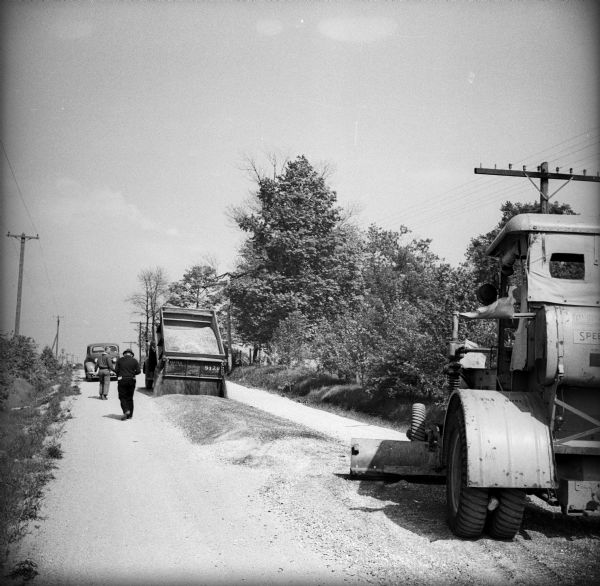 A dump truck full of loose gravel is emptying its load onto a street. A leveler is following behind to redistribute the gravel evenly across the road. There are two men walking to the left of the dump truck. The back of the dump truck has text that reads "WPA 5129."